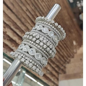 May include: A collection of silver bangles adorned with clear rhinestones. The bangles, of different widths, showcase detailed patterns and large, circular central stones. The jewellery is presented on a silver rod, set against a blurred wooden backdrop.