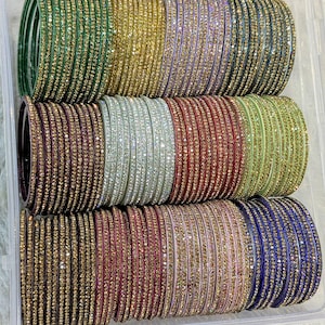Puede incluir: Un recipiente de plástico transparente lleno de pilas de pulseras coloridas. Las pulseras son de varios colores, incluyendo verde, dorado, morado, marrón, plateado, rojo, verde claro, negro, rosa y azul. Cada pulsera está adornada con pequeños adornos brillantes.