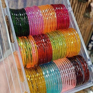May include: A clear plastic container filled with stacks of colorful glass bangles. The bangles are in various colors, including green, pink, orange, red, and blue. The bangles are stacked neatly inside the container, ready for sale.