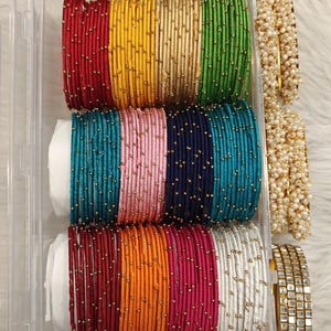 Puede incluir: Una colección de pulseras coloridas en un recipiente de plástico transparente. Las pulseras son de varios colores, incluyendo rojo, dorado, verde, turquesa, rosa, azul marino, naranja, magenta y blanco. Algunas pulseras están adornadas con pequeñas cuentas doradas. También se ven pulseras adicionales con perlas y detalles dorados cuadrados.