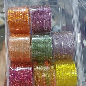 May include: A clear plastic container holding nine stacks of colourful bangles. The bangles are in shades of orange, purple, green, pink, yellow, and orange with gold glitter. The container is open.