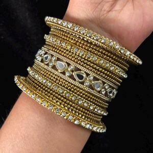 Op de afbeelding: Een stapel goudkleurige armbanden met een textuurrijke metalen afwerking en ingelegde heldere kristallen. De set bevat verschillende dunne, kraalachtige banden en een bredere centrale armband versierd met druppelvormige spiegelaccenten.