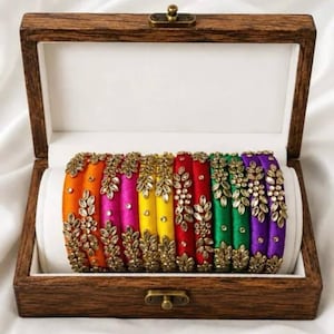 May include: A set of multi-colored silk thread bangles arranged in a wooden box. The bangles feature vibrant orange, pink, yellow, red, green, and purple colors, each adorned with a delicate gold-toned leaf pattern embellished with small clear stones.