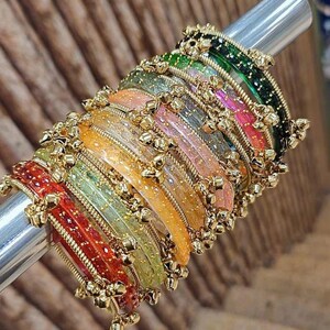 May include: A selection of vibrant bangles in red, orange, yellow, green, pink, and green. Each bangle features gold-coloured detailing and small dangling charms. The bangles are presented on a silver rod against a textured wooden backdrop.