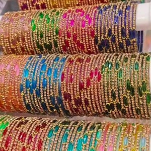 Puede incluir: Una colección de brazaletes metálicos delgados dispuestos en cuatro filas horizontales. Cada brazalete presenta un acabado dorado metálico con pequeñas piedras transparentes incrustadas y detalles ovalados de colores en tonos rojo, amarillo, verde, azul y rosa.
