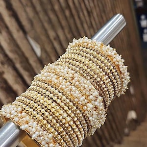 Puede incluir: Una colección de pulseras doradas adornadas con pequeñas perlas blancas. Las pulseras están apiladas en una varilla plateada, mostrando su intrincado diseño y textura. Las joyas se presentan sobre un fondo de madera borroso.