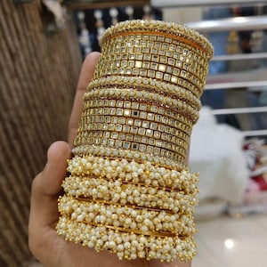 May include: A stack of gold-colored bangles. Some bangles feature a square, mirrored design, while others are adorned with small, white, pearl-like beads. The bangles are held in a hand, showcasing the jewelry's intricate details and craftsmanship.
