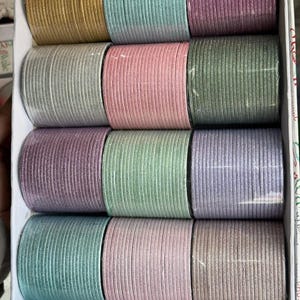 Puede incluir: Una colección de doce juegos de brazaletes finos y texturizados dispuestos en una caja blanca. Los brazaletes presentan un acabado brillante en varios colores, incluyendo oro, azul claro, morado, plata, rosa, verde oscuro, lavanda y marrón claro.