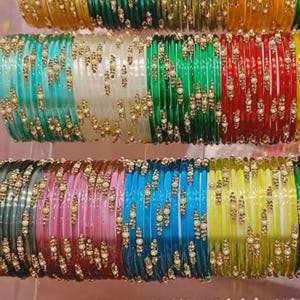 Puede incluir: Filas de pulseras de cristal de colores en tonos amarillo, verde, rojo, rosa, azul y dorado. Cada pulsera está adornada con pequeños adornos dorados. Las pulseras están apiladas, mostrando una variedad de colores y diseños.