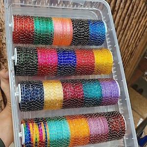 Könnte beinhalten: Ein durchsichtiger Kunststoffbehälter gefüllt mit bunten Glasarmreifen. Die Armreifen sind in Reihen angeordnet und zeigen eine Vielzahl von Farben, darunter Rot, Grün, Pink, Blau und Gold. Die Armreifen sind wahrscheinlich aus Glas und eine traditionelle Schmuckform.