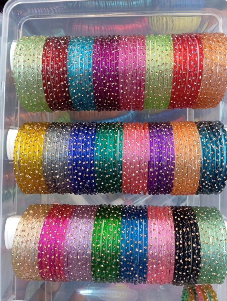 A collection of colorful bangles in a clear plastic container. The bangles are arranged in rows, showcasing a variety of colors including green, red, blue, purple, and gold. Each bangle is adorned with small, sparkling embellishments.