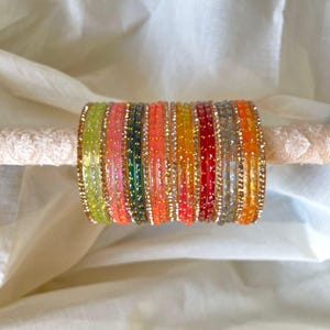 May include: A collection of colorful bangles in shades of green, pink, orange, and red, adorned with gold-colored accents. The bangles are displayed on a light-colored, textured surface, creating a vibrant and eye-catching display. These accessories are perfect for adding a pop of color.