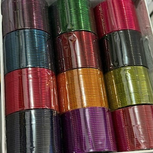 Könnte beinhalten: Eine Schachtel gefüllt mit Stapeln bunter Armreifen. Die Armreifen sind in verschiedenen Farben erhältlich, darunter Rot, Grün, Gold, Lila und Schwarz. Die Armreifen bestehen aus einem glänzenden Material und sind ordentlich in Reihen angeordnet.