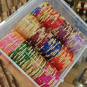 Puede incluir: Un recipiente de almacenamiento de plástico transparente lleno de varias pilas de brazaletes coloridos. Cada brazalete presenta un acabado vibrante y translúcido acentuado con pequeñas cuentas metálicas doradas en un patrón repetitivo.