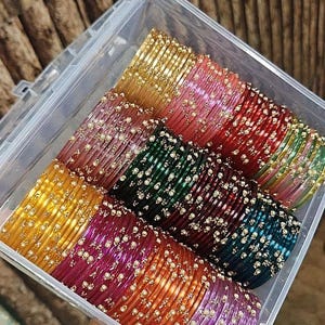 May include: A clear plastic box containing rows of colourful bangles. The bangles are in shades of gold, pink, red, green, and purple. Each bangle is decorated with small, white embellishments, creating a shimmering effect.