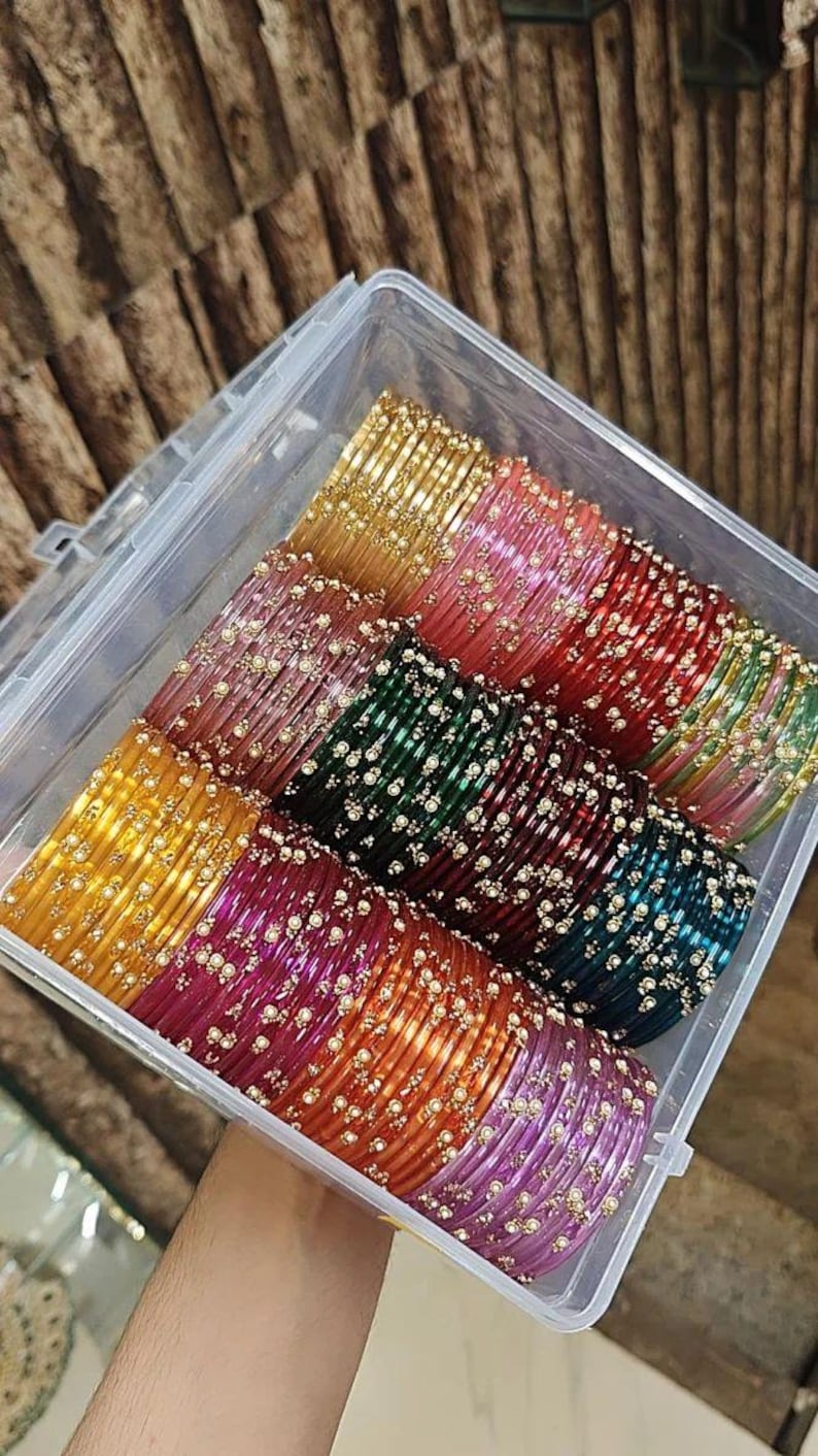 May include: A clear plastic box containing rows of colourful bangles. The bangles are in shades of gold, pink, red, green, and purple. Each bangle is decorated with small, white embellishments, creating a shimmering effect.