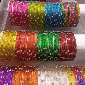 May include: A collection of colorful glass bangles, each adorned with small, sparkling embellishments. The bangles are arranged in stacks, showcasing a variety of vibrant hues including pink, green, yellow, and blue. The decorative bangles are displayed on white supports.