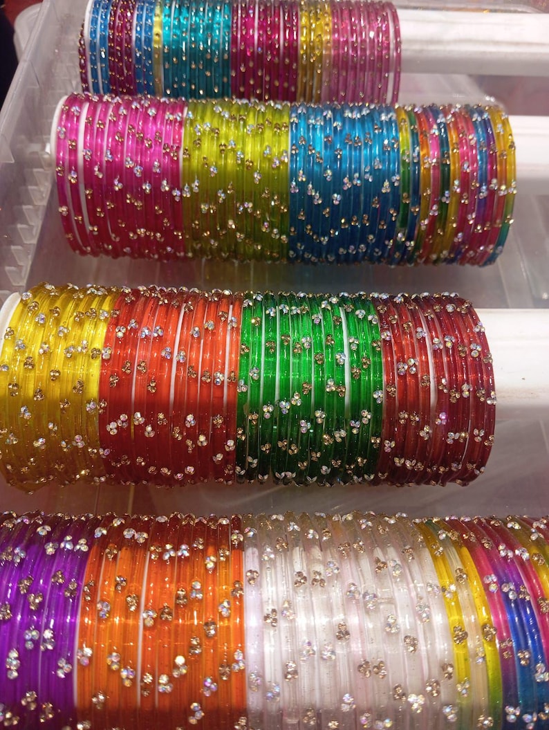 May include: A collection of colorful glass bangles, each adorned with small, sparkling embellishments. The bangles are arranged in stacks, showcasing a variety of vibrant hues including pink, green, yellow, and blue. The decorative bangles are displayed on white supports.
