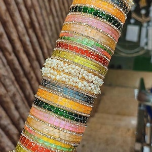 Puede incluir: Una pila de pulseras coloridas en varios tonos, incluyendo amarillo, verde, rosa y rojo. Cada pulsera está adornada con pequeñas perlas blancas y detalles dorados. Las pulseras están apiladas, creando una exhibición vibrante y llamativa.