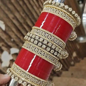 Puede incluir: Una pila de brazaletes rojos con adornos dorados y gemas transparentes. Los brazaletes se sostienen sobre una varilla plateada. Las bandas doradas están decoradas con motivos florales. Accesorio tradicional indio.