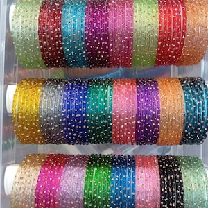 May include: A collection of colorful bangles in a clear plastic container. The bangles are arranged in rows, showcasing a variety of colors including green, red, blue, purple, and gold. Each bangle is adorned with small, sparkling embellishments.