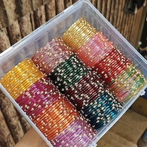 May include: A clear plastic container filled with colourful bangles. The bangles are in shades of gold, pink, red, green, and purple, with small gold accents. The bangles are stacked neatly in rows.