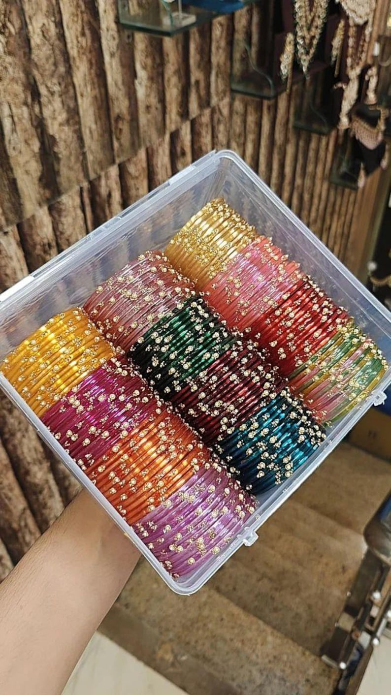 May include: A clear plastic container filled with colourful bangles. The bangles are in shades of gold, pink, red, green, and purple, with small gold accents. The bangles are stacked neatly in rows.