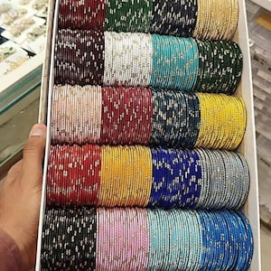 May include: A box filled with stacks of colorful bangles. The bangles come in various colors, including red, green, gold, blue, and pink. Each bangle has a metallic sheen and is adorned with a delicate pattern.