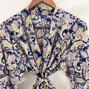 May include: A navy blue paisley patterned robe hanging on a wooden hanger. The robe features a repeating design of pink, yellow, and white paisley shapes. The robe is tied at the waist with a matching fabric tie.