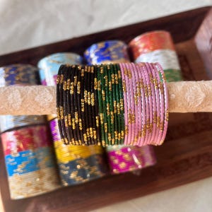 May include: A collection of colorful bangles in a wooden tray. The bangles are in various colors, including black, green, pink, and gold. The bangles are stacked together, and some have a glittery finish. The bangles are a traditional type of jewelry.