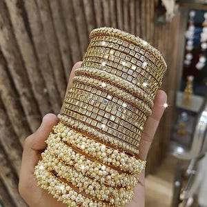 May include: A stack of gold-colored bangles with square-cut mirrored accents and a row of small, white pearls. The bangles are stacked on top of each other, creating a layered effect. The jewelry is displayed in a store setting.