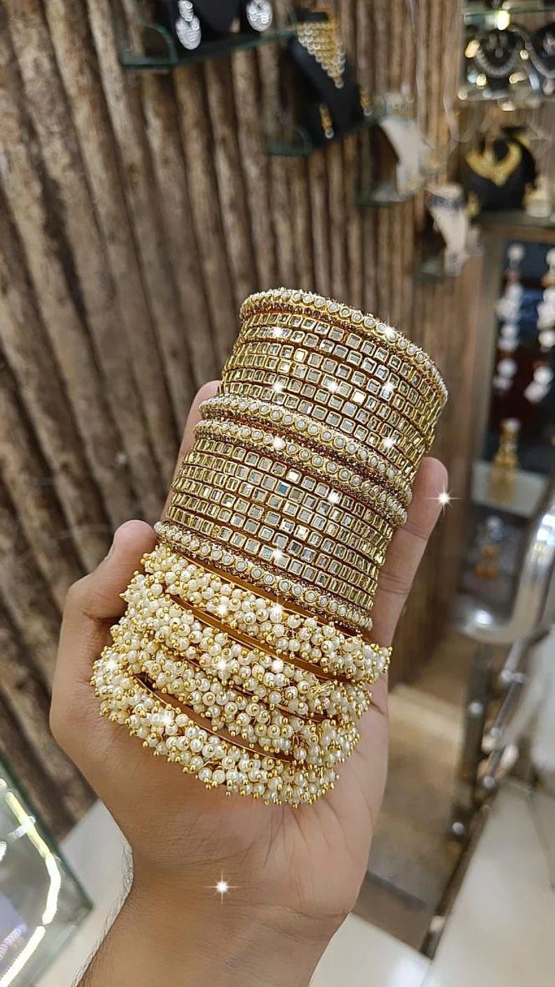 May include: A stack of gold-colored bangles with square-cut mirrored accents and a row of small, white pearls. The bangles are stacked on top of each other, creating a layered effect. The jewelry is displayed in a store setting.