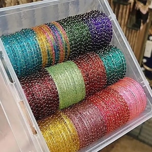 Puede incluir: Una caja de almacenamiento de plástico transparente que contiene varias pilas de brazaletes finos y redondos en colores vibrantes como turquesa, amarillo, morado, verde, rojo, lima, granate, rosa y dorado. Cada brazalete tiene una textura brillante.
