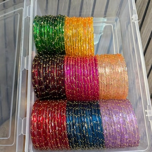 Puede incluir: Un recipiente de plástico transparente lleno de pilas de pulseras de cristal de colores. Las pulseras son en tonos de verde, amarillo, marrón, rosa, naranja, rojo, azul y morado, con detalles dorados. Las pulseras están apiladas ordenadamente dentro del recipiente.