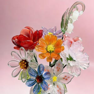 Peut inclure: Un bouquet de fleurs en verre coloré dans un vase transparent. L'arrangement comprend des fleurs rouges, oranges, bleues, blanches et roses avec des tiges et des feuilles vertes. Le fond est rose clair.