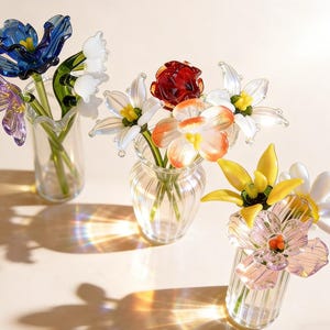 Peut inclure: Trois petits vases en verre contiennent chacun un bouquet de fleurs en verre colorées. Les fleurs comprennent des tulipes bleues, des lys blancs, une rose rouge et des marguerites jaunes et roses. Les vases sont transparents et côtelés.