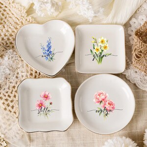 Può includere: Quattro piattini in ceramica bianca con bordi dorati. Il piattino a forma di cuore presenta fiori blu e il nome "aurora". Il piattino quadrato mostra narcisi gialli e "grace". Il piattino quadrato smerlato ha fiori rosa e "stella". Il piattino rotondo mostra peonie rosa e "evelyn".