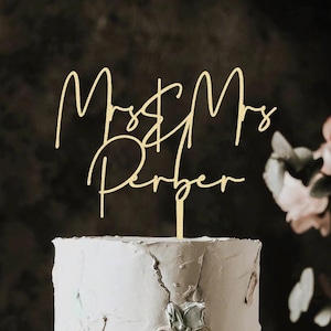 Op de afbeelding: Een goudkleurige taarttopper met de woorden "Mr & Mrs Perber" in een cursief lettertype. De topper staat op een witte taart met getextureerde frosting. Roze bloemen staan op de achtergrond.