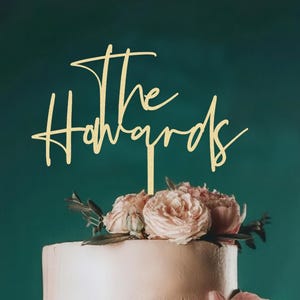 May include: A gold cake topper with the words "The Howards" in cursive script. The cake is decorated with pink roses and greenery. The background is a dark teal colour. The cake is frosted white.