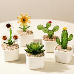Peut inclure: Plusieurs sculptures de plantes en verre, dont un tournesol, des marguerites, un cactus et une succulente, chacune dans un petit pot carré blanc rempli de gravier. Les plantes en verre sont de différentes nuances de vert, jaune et marron.