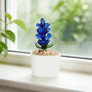 Peut inclure: Une fleur décorative en verre dans un pot carré blanc. La fleur est d'un bleu vif avec des feuilles vertes, et le pot est rempli de petites pierres de couleur claire. La plante est sur un rebord de fenêtre blanc avec un arrière-plan vert flou.