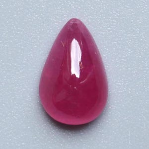 May include: A polished, teardrop-shaped ruby gemstone. The gem is a deep, saturated red color with a smooth, reflective surface. The stone is set against a light gray background.