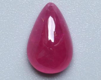 Puede incluir: Una gema de rubí pulida en forma de lágrima. La gema es de color rojo intenso y saturado, con una superficie lisa y reflectante. La piedra se encuentra sobre un fondo gris claro.
