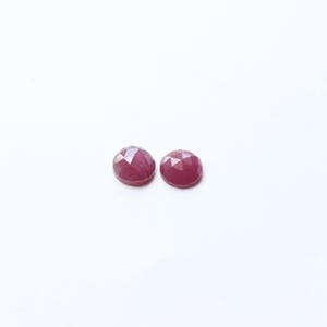 May include: Two faceted, round, deep red gemstones. The gems have a polished, reflective surface and are set against a plain white background. These could be used for jewelry making or as decorative elements.