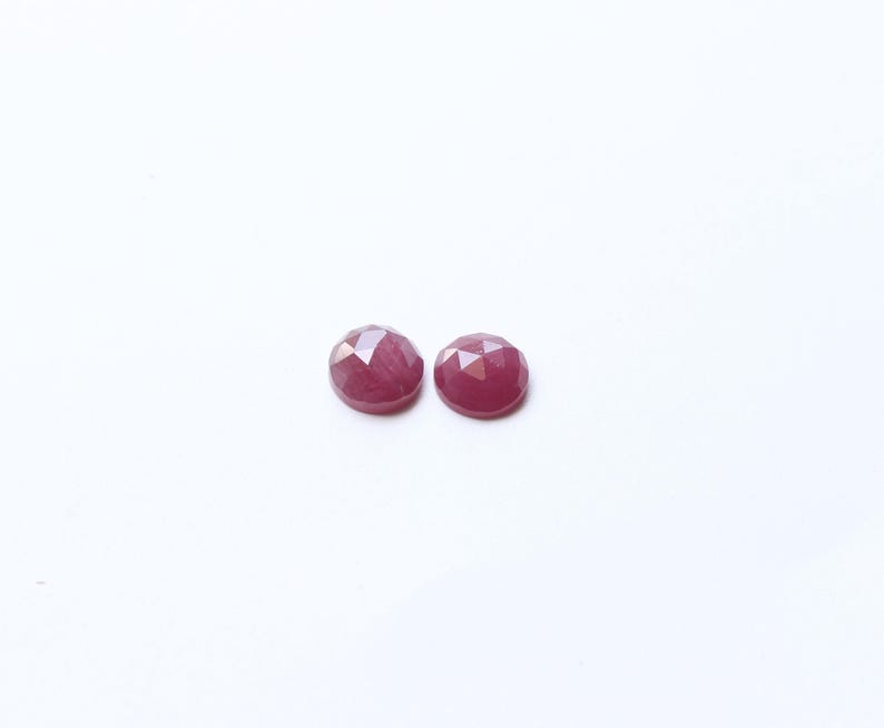 May include: Two faceted, round, deep red gemstones. The gems have a polished, reflective surface and are set against a plain white background. These could be used for jewelry making or as decorative elements.