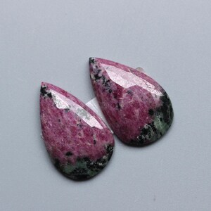 Könnte beinhalten: Zwei tropfenförmige Edelsteine mit facettierter Oberseite. Die Steine sind eine Mischung aus tiefem Rosa, Schwarz und Grün. Die Edelsteine liegen vor einem hellgrauen Hintergrund.