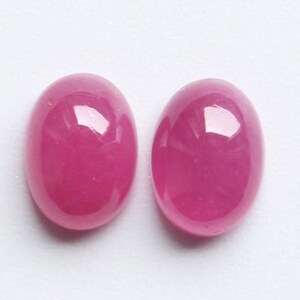 Könnte beinhalten: Zwei ovale, polierte Edelsteine in einem leuchtenden Magenta-Farbton. Die Steine haben eine glatte, glänzende Oberfläche, die das Licht reflektiert. Die Edelsteine sind vor einem schlichten weißen Hintergrund platziert. Ideal für Schmuckherstellung.