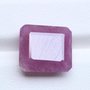 May include: A rectangular, faceted pink gemstone with a polished surface. The stone features a clear, light-reflecting centre facet surrounded by a deeper pink, opaque border with visible internal inclusions and a smooth, geometric cut.