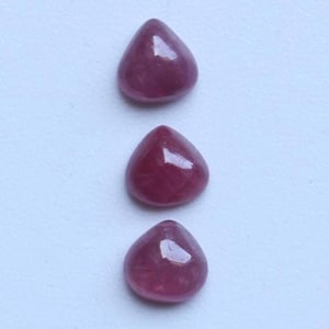 May include: Three polished, teardrop-shaped gemstones in a deep magenta colour. The gems are arranged vertically against a light background. Each stone has a smooth, reflective surface, catching the light.