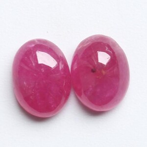 May include: Two oval-shaped, polished gemstones in a deep pink colour. The gems have a smooth, reflective surface, with subtle internal patterns. The gemstones are set against a plain white background.